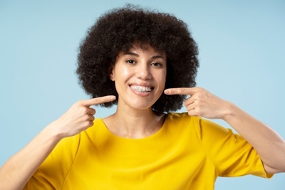 Woman smiling and pointing at their teeth, wearing a yellow shirt against a blue background.